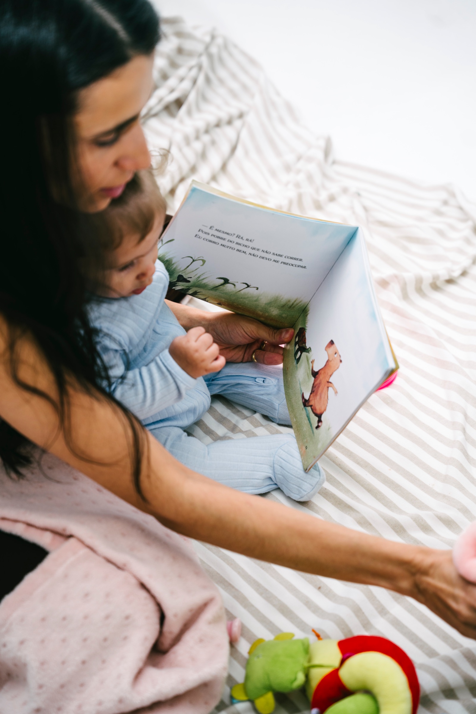 Uma mulher de cabelos escuros está sentada sobre uma cama com lençol listrado em tons claros, segurando um bebê vestido com um macacão azul-claro no colo. Juntos, observam um livro infantil aberto, apoiado nas mãos da mulher. A página visível traz uma ilustração de um animal e um pequeno trecho de texto. O bebê olha atentamente para o livro, com uma das mãos levemente levantada, demonstrando interesse pela leitura. Ao redor, há uma manta clara ao fundo e um brinquedo de pelúcia colorido próximo aos pés. A cena transmite um momento de carinho, conexão e leitura compartilhada entre adulto e criança, em um ambiente acolhedor e iluminado.
